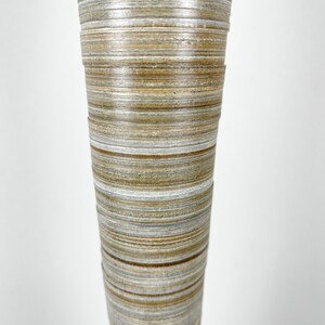 May include: A tall, cylindrical vase featuring a layered, horizontal striped pattern in shades of tan, brown, gray, and white. The surface has a textured, stacked appearance with visible rings that wrap around the entire exterior of the object.