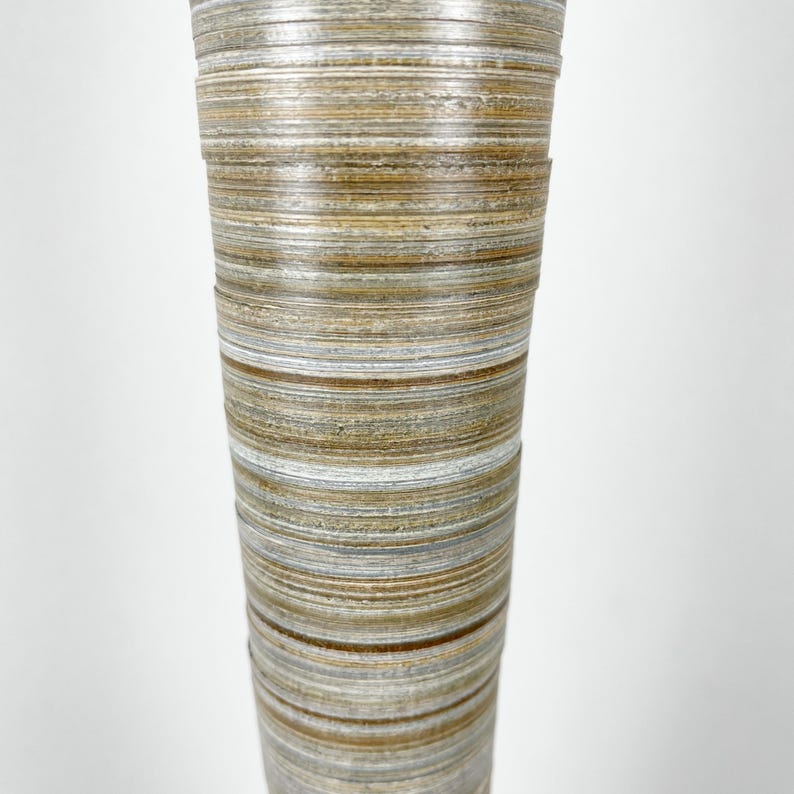 May include: A tall, cylindrical vase featuring a layered, horizontal striped pattern in shades of tan, brown, gray, and white. The surface has a textured, stacked appearance with visible rings that wrap around the entire exterior of the object.