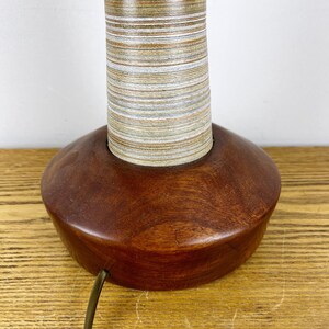 May include: A table lamp base featuring a smooth, dark brown wooden circular foundation and a cylindrical neck wrapped in tightly wound, multi-colored horizontal thread in shades of tan, beige, white, and muted green with a power cord.