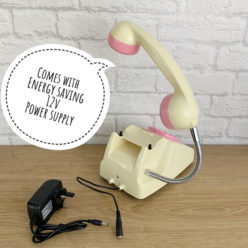 May include: A cream-colored rotary phone with a pink handset and a flexible gooseneck. The phone is connected to a black power adapter with a UK plug. The text "COMES WITH ENERGY SAVING 12V POWER SUPPLY" is in a speech bubble.