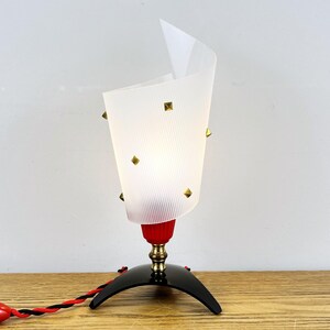 May include: A vintage table lamp with a white, ribbed shade adorned with gold-colored square studs. The lamp features a red base, a brass-colored stem, and a black, curved base. The electrical cord is red and black.