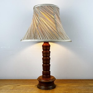 May include: A table lamp with a turned wooden base featuring a dark brown finish and a stacked cylindrical design. The lamp is topped with a flared, bell-shaped lampshade covered in a fine, multi-colored striped fabric in shades of tan and grey.