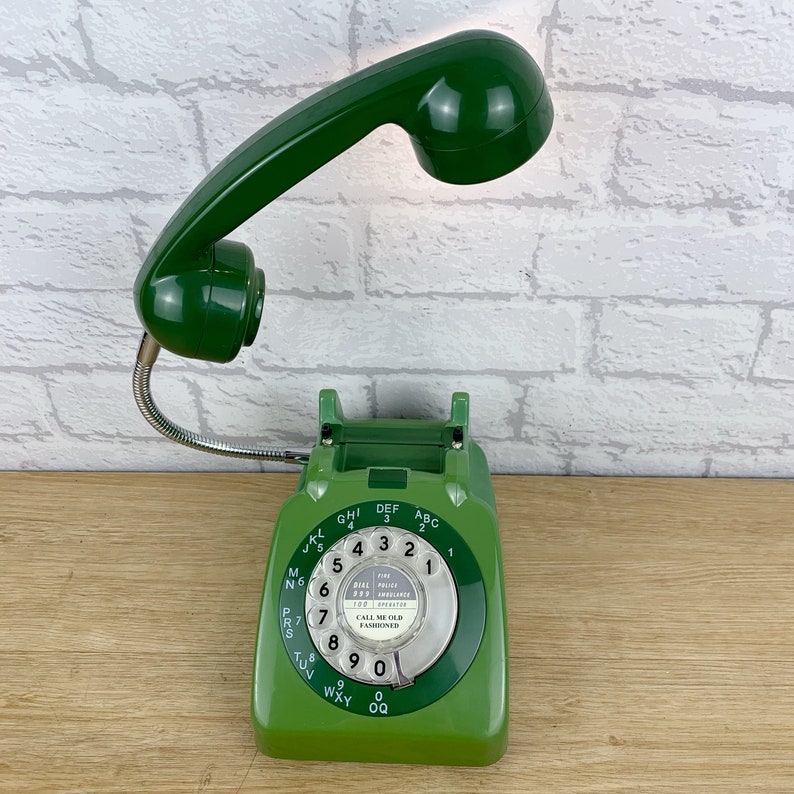 May include: A green rotary dial phone with a green handset attached to a flexible arm. The phone has a white dial with black numbers and the text "DIAL 999 FOR POLICE FIRE AMBULANCE 100 FOR OPERATOR" and "CALL ME OLD FASHIONED".