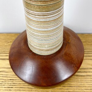 May include: A cylindrical object featuring a base made of smooth, dark brown polished wood. Rising from the center is a tall, narrow column composed of tightly stacked, thin layers of material in alternating shades of tan, white, and blue.