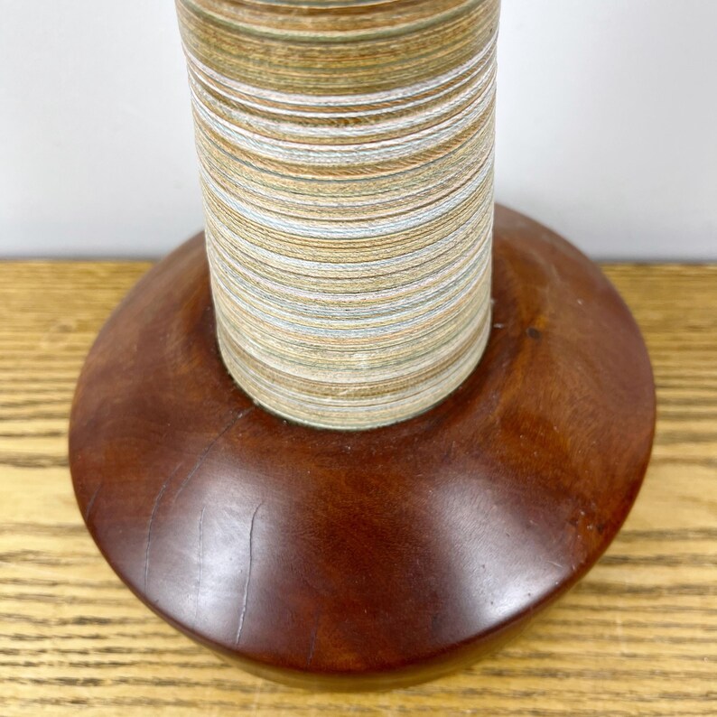 May include: A cylindrical object featuring a base made of smooth, dark brown polished wood. Rising from the center is a tall, narrow column composed of tightly stacked, thin layers of material in alternating shades of tan, white, and blue.