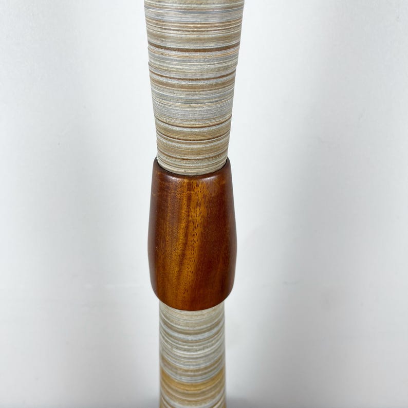 May include: A tall, cylindrical lamp base featuring a central section of polished dark brown wood. The upper and lower portions are composed of stacked, layered material with horizontal bands of beige, gray, and white tones.