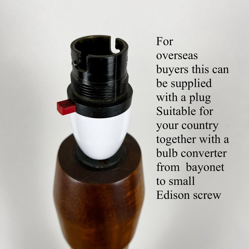 May include: A wooden lamp base featuring a black and white bayonet light socket with a small red switch. The text reads: For overseas buyers this can be supplied with a plug suitable for your country together with a bulb converter from bayonet to small Edison screw.