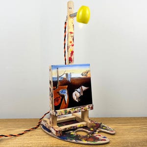 May include: A unique desk lamp featuring a small canvas with a surrealist painting, mounted on a wooden easel. The lamp has a yellow shade and a wooden post with a decorative cord. The base is a painted artist's palette.