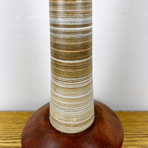 May include: A table lamp base featuring a cylindrical column with horizontal layers of brown, tan, and white wood grain patterns, set upon a smooth, rounded, dark reddish-brown wooden base with a polished finish and visible wood grain.