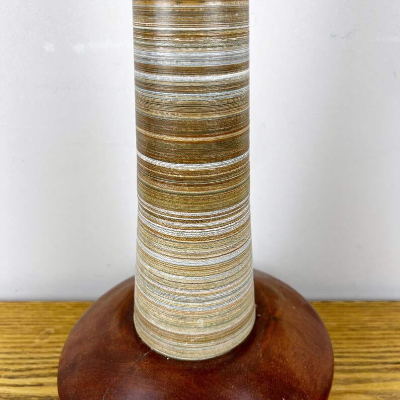 May include: A table lamp base featuring a cylindrical column with horizontal layers of brown, tan, and white wood grain patterns, set upon a smooth, rounded, dark reddish-brown wooden base with a polished finish and visible wood grain.