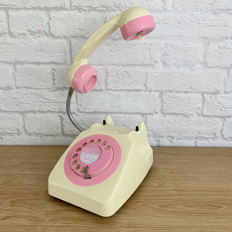 May include: A vintage-style rotary phone with a cream-colored base and a pink rotary dial. The phone has a flexible pink handset with a cream-colored base. The phone has a pink dial with the text "CALL ME UP,  FANNY LOVE" printed on it.