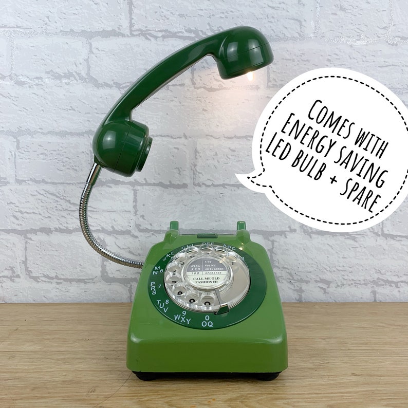 May include: Green rotary dial phone with a green phone handset attached to a flexible gooseneck lamp. The lamp has a white LED bulb and is turned on. The phone has the text "CALL ME OLD FASHIONED" on the dial. The phone is on a wooden surface. The text "COMES WITH ENERGY SAVING LED BULB + SPARE" is in a speech bubble.
