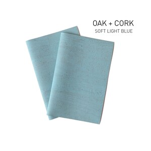 Op de afbeelding: Twee vellen lichtblauw kurkmateriaal met een subtiele gespikkelde textuur. De tekst "OAK + CORK SOFT LIGHT BLUE" is boven de vellen gedrukt.
