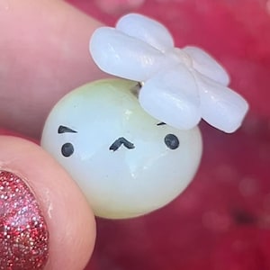 May include: A small, round, light green and white bead with a cartoon face and a white flower on top. The bead has black eyes and a curved mouth. The background is a blurred red.