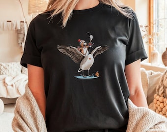 Puede incluir: Una camiseta negra con un gráfico de un oso de dibujos animados con un gorro de Papá Noel montando un ganso mientras sostiene un palo de hockey. El ganso lleva una bandera canadiense.
