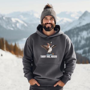 May include: A dark grey hooded sweatshirt featuring a graphic of a goose riding a smaller goose with a hockey stick. The text "Tariff This, Hoser!" is printed below the graphic.