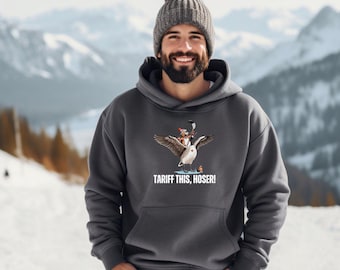 Puede incluir: Una sudadera con capucha gris oscuro con un gráfico de un ganso montando a otro ganso más pequeño con un palo de hockey. El texto "Tariff This, Hoser!" está impreso debajo del gráfico.