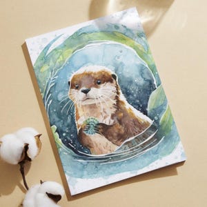 Puede incluir: Ilustración en acuarela de una nutria sosteniendo una concha, rodeada de plantas acuáticas. La nutria tiene pelaje marrón y cara blanca, sobre un fondo azul y verde. La obra de arte está en una tarjeta rectangular.