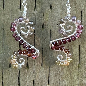 Peut inclure: Une paire de boucles d'oreilles en argent avec un motif en spirale composé de petites pierres précieuses colorées. Les pierres précieuses sont dans des tons de rose, rouge, brun et jaune.