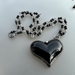 Op de afbeelding: Een ketting met een zwarte hartvormige hanger, een zilveren ketting en zwarte kralen. De hanger is gemaakt van een zwarte steen met een zilveren frame.