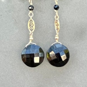 May include: A pair of gold dangle earrings with black faceted gemstone drops. The earrings feature a delicate gold chain with a small black bead and a decorative gold charm.