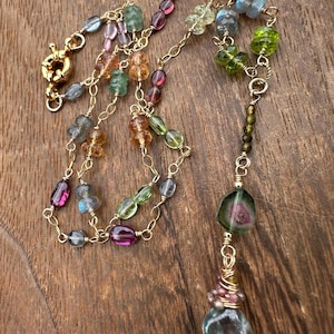 Può includere: Una collana a catena dorata con una varietà di perline di pietre preziose sfaccettate multicolori nei toni del rosa, verde, blu e ambra. La collana include una grande fetta di tormalina anguria centrale e un pendente a goccia blu.