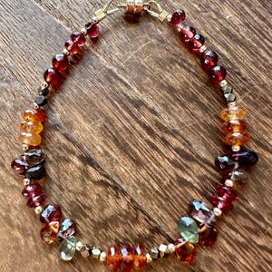 May include: A beaded bracelet featuring a variety of gemstones in shades of red, orange, and green. The bracelet includes faceted and round beads, with gold-colored metal accents and a magnetic clasp. The bracelet is displayed on a wooden surface.