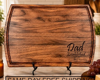 May include: A rectangular dark brown wooden cutting board with rounded corners and a juice groove along the perimeter. The board features the engraved text "Dad Established 10.31.00 04.14.04" in the lower right corner. It is displayed on a black metal stand.