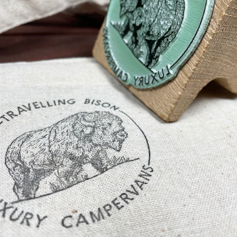 Pu&ograve; includere: Una borsa di tela bianca con un'illustrazione in bianco e nero di un bisonte. Il testo "Travelling Bison Luxury Campervans" &egrave; stampato intorno al bisonte.