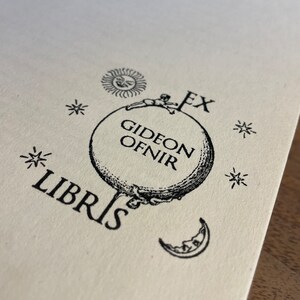 May include: Black and white illustration of a globe with a figure sitting on top. The globe has the text "Gideon Ofnir" on it. The illustration is surrounded by stars and a sun and moon.
