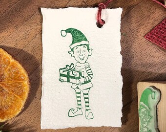 May include: A green and white Christmas gift tag featuring a cartoon elf holding a gift box. The elf is wearing a green hat with a white pom-pom and a striped shirt.