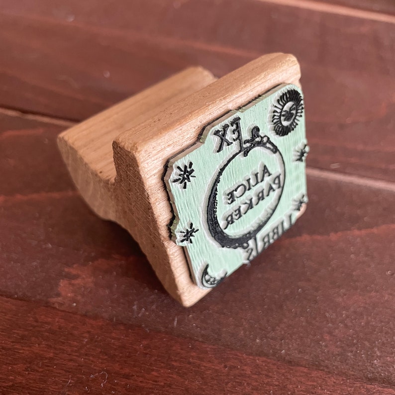 May include: A wooden stamp with a green and black design. The stamp features the text "Alice Parker" in a circle with a crescent moon and stars. The stamp is labeled "Ex Libris".