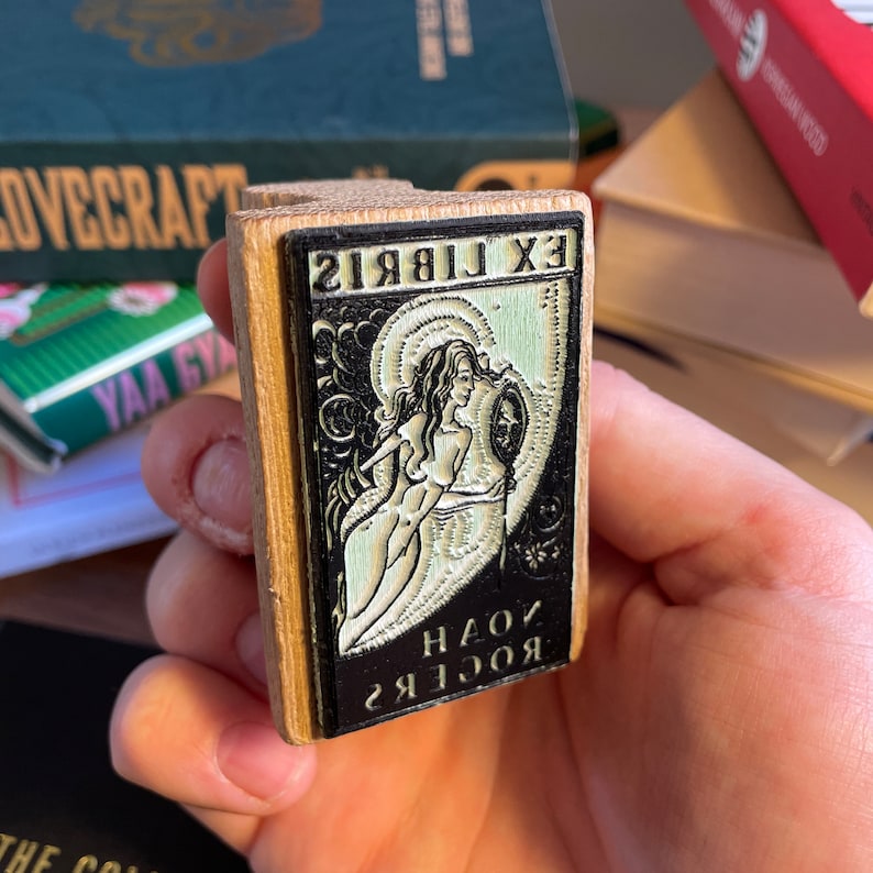 Pu&ograve; includere: Un timbro in legno con un'immagine in bianco e nero di una donna che tiene uno specchio. Il testo sul timbro recita "EX LIBRIS" e "ЯОСЯ К ЯОСЯ".