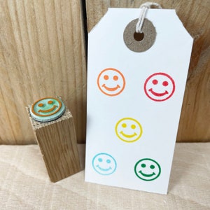 May include: A set of six colourful rubber stamps featuring a smiley face design. The stamps are arranged in a circle on a white tag with a hole in the top. The stamps are red, orange, yellow, green, blue, and purple.