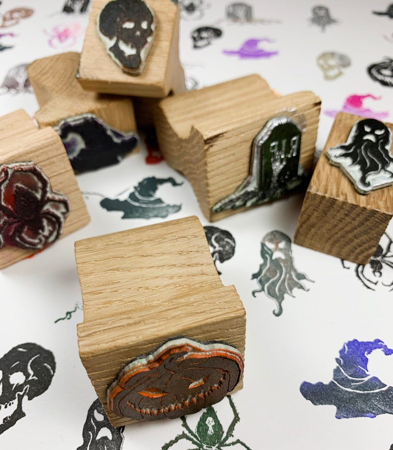 Pu&ograve; includere: Una collezione di timbri in legno a tema Halloween, con disegni come un teschio, un ragno, un fantasma, una lapide e una zucca. I timbri hanno una finitura in legno naturale e sono disposti su una superficie bianca con motivi di Halloween stampati.