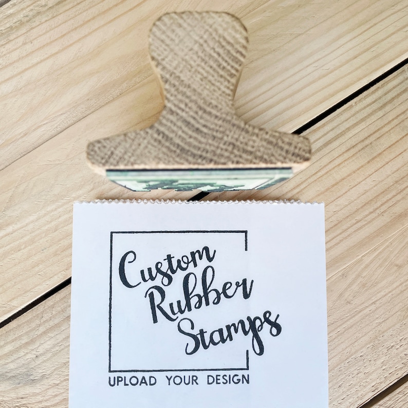 Pu&ograve; includere: Un timbro in gomma di legno con sfondo bianco e testo nero che recita "Custom Rubber Stamps" in una cornice quadrata. Sotto la cornice, il testo "UPLOAD YOUR DESIGN" &egrave; stampato in nero.