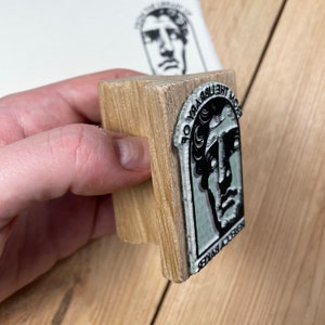 May include: A wooden stamp with a black and white image of a man's face. The text "FROM THE LIBRARY OF" is printed around the image.