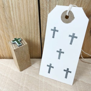Puede incluir: Un bloque de madera con un pequeño sello de cruz verde y blanco en la parte superior. Una etiqueta blanca con cinco cruces grises impresas en ella.