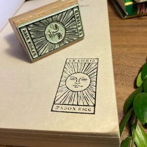 Może przedstawiać: Drewniana pieczątka z motywem słońca i napisem "EX LIBRIS JASON RIGG" odbitym na książce. Pieczątka ma wz&oacute;r twarzy słońca z promienistymi liniami. Pieczątka znajduje się obok odbitego obrazu na książce.
