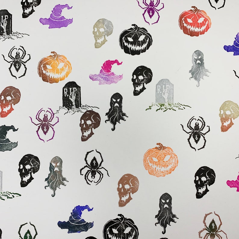 Pu&ograve; includere: Un motivo ripetuto di illustrazioni a tema Halloween su sfondo bianco. Il design include zucche intagliate, teschi, ragni, cappelli da strega, lapidi con il testo "RIP" e fantasmi in vari colori.