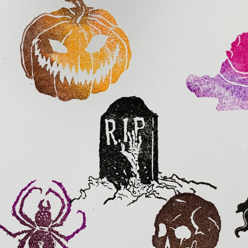 Pu&ograve; includere: Stampa a tema Halloween con una zucca intagliata, una lapide con "R.I.P." e una mano scheletrica, un ragno e un teschio. La zucca &egrave; arancione e marrone, la lapide &egrave; nera e il ragno &egrave; viola.