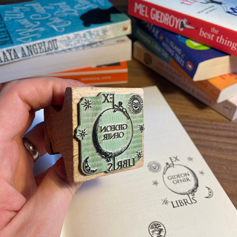 May include: A wooden stamp with a green and black design. The stamp features a globe with the text "GIDEON OFNIR" and "LIBRIS" around it. The stamp also has a sun and a crescent moon.