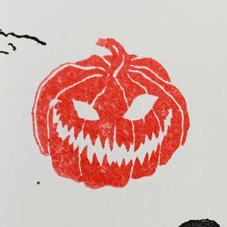 Pu&ograve; includere: Illustrazione di una zucca rossa intagliata con un sorriso minaccioso e occhi triangolari. Il design a tema Halloween &egrave; stampato su sfondo bianco. La zucca ha un aspetto strutturato, che suggerisce una stampa o un timbro.