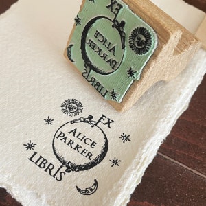May include: A wooden stamp with a green ink pad featuring the text "Alice Parker" and "Libris" surrounded by stars and a crescent moon. The stamp is being used to create an imprint on a white surface with the text "Alice Parker", "Libris", and "Ex" surrounded by stars and a crescent moon.