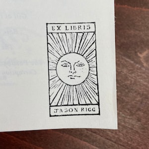 Może przedstawiać: Czarno-biały ekslibris z napisem "EX LIBRIS" u g&oacute;ry i "JASON RIGG" u dołu. W środku znajduje się słońce z twarzą i promieniami.