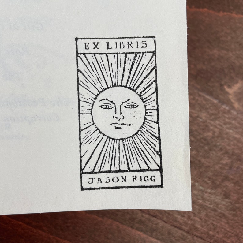 Może przedstawiać: Czarno-biały ekslibris z napisem "EX LIBRIS" u g&oacute;ry i "JASON RIGG" u dołu. W środku znajduje się słońce z twarzą i promieniami.