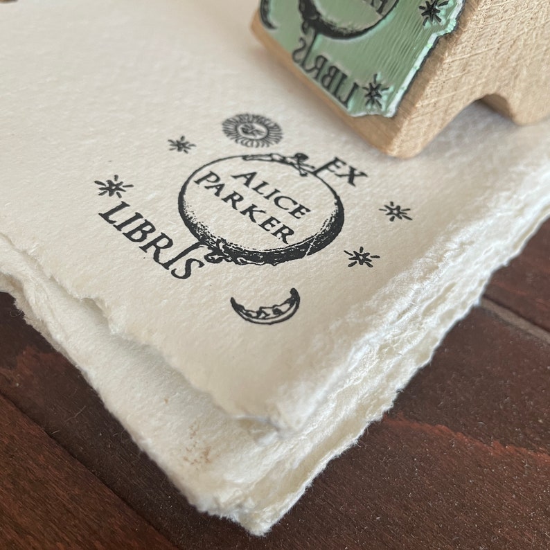 May include: A black and white stamp with the text "Alice Parker Ex Libris" and a circular design with a skull and crossbones. The stamp is on a white paper background.