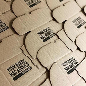 Pu&ograve; includere: Primo piano di pi&ugrave; scatole di cartone con la scritta "Your Scents of Humour Has Arrived" stampata su di esse. Ogni scatola ha un piccolo rettangolo nero con la scritta "Tin Opener" stampata su di esso.