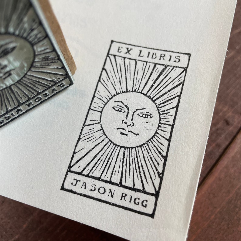 Może przedstawiać: Czarno-biały wz&oacute;r promieni słonecznych z twarzą, odbity na białym papierze. Na stemplu widnieje napis "EX LIBRIS" u g&oacute;ry i "JASON RIGG" u dołu. Widoczny jest metalowy stempel z tym samym wzorem promieni słonecznych.