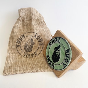Pu&ograve; includere: Un timbro in legno con un design verde e nero di una creatura con il testo "YOUR LOGO HERE" attorno al bordo. Il timbro &egrave; su una base in legno e viene fornito con un sacchetto di juta.